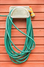 Green plastic garden hose and nozzle hanging on the wall of a red wooden house