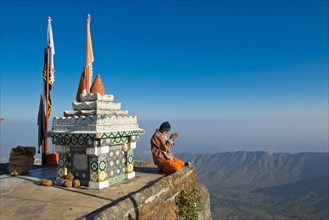 Sadhu or holy man sitting with his hands folded in prayer on the precipice in front of a Hindu temple shrine with prayer flags