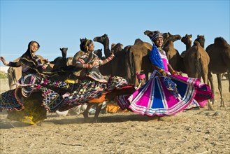 Three young women in colourful dresses dancing in front of a herd of camels