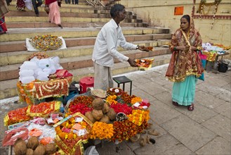 Man at a stall offering small bowls with flowers and ritual offerings to a passing woman
