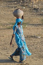 Indian woman wearing a light blue sari carrying a clay water jug on her head