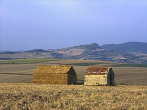 Bales of straw stacked in the shape of a house next to a little stone house