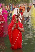 Woman wearing a red sari with offerings standing in water during the Hindu Chhath Festival