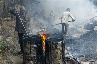 A worker heating tar in barrels with an open fire at a road construction site