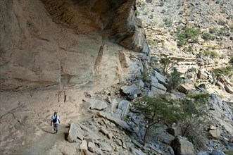 Local hiker walking under a rock overhang on the path to the abandoned settlement of Sap Bani Khamis on the western flank of the Grand Canyon of Oman