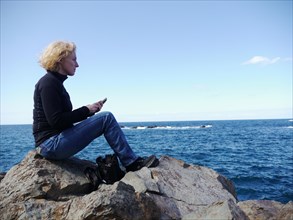 Woman with phone sitting on a rock and looking over the sea