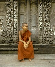 Monk sitting in front of a wooden door with teak carvings