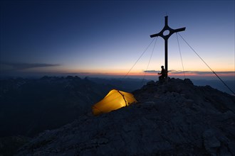 Mountaineer with a tent at the summit cross on Großer Krottenkopf Mountain at dusk