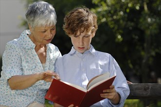 Grandmother and grandson are looking at a red book together on park bench