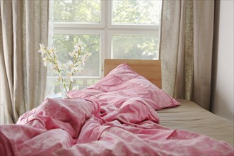 A crumpled bed with a pink blanket by a window