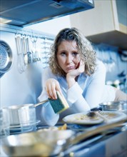 Annoyed woman holding a sponge while standing in a kitchen with dirty dishes