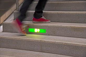 Person rushing up a public staircase with a green illuminated emergency exit sign
