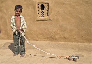Boy playing with a home-made toy car outside a mud house