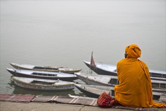 Brahmin or Hindu priest sitting on a blanket on the banks of the Ganges River with several boats tied to the shore