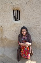 Smiling girl wearing a headscarf and traditional clothing sitting in front of a house wall with a window opening