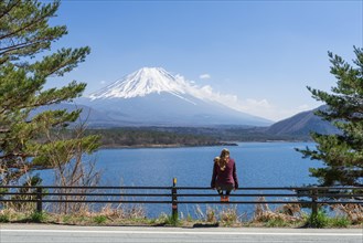 Young woman sitting on a railing next to a road and looking across the lake to the volcano Mt Fuji