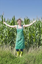 Young woman in work clothes standing with outstretched arms in front of a maize field