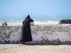A Berber with black djellaba leaning against the harbor wall