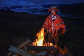 Indio man with hat and colorful traditional poncho standing at a campfire in the Andes