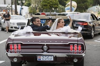 Bride and groom riding in an open Ford Mustang convertible car through the city
