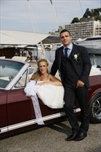 Bride and groom posing with an open Ford Mustang convertible car at a harbour
