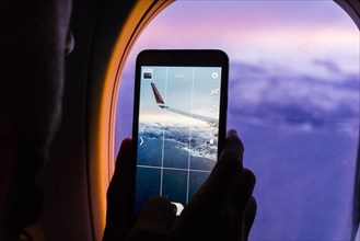 Silhouette of a man taking photos of the mountains and airplane wing using a smartphone from an airplane window