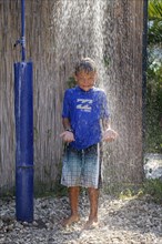 5-year-old boy wearing a blue t-shirt standing under a running shower in a garden