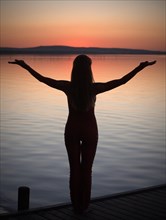 Woman with backlighting doing a yoga pose on a wooden pier at a lake after sunset
