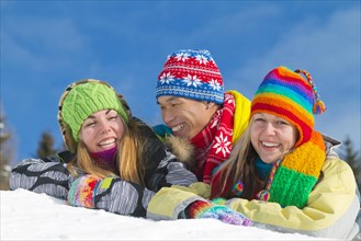 Young Asian man and two young European women smiling while lying on a mound of snow and wearing colourful winter clothing