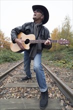 Elderly man wearing jeans and a black leather jacket with a black cowboy hat and a western guitar singing on a train track
