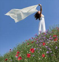 Young woman wearing a white summer dress on the slope of a flowering meadow while holding a white silk scarf fluttering in the wind