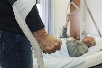 Elderly woman standing on a crutch in front of an elderly man lying in a hospital bed