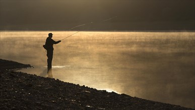 A fly fisherman standing on the shore of the Obernau reservoir at sunrise