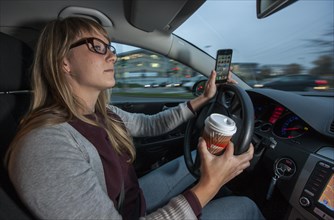 Young woman driving a car in the city centre while holding her cell phone and a cup of coffee at the same time
