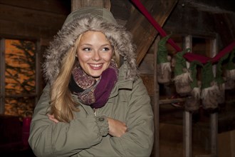 Smiling young woman wearing a green parka standing with her arms crossed in front of a hiking hut decorated for Christmas