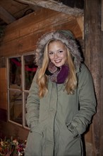 Smiling young woman wearing a green parka standing in front of a hiking hut decorated for Christmas