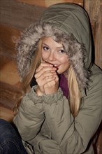 Young woman wearing a green parka sitting in front of a hiking hut and warming her hands