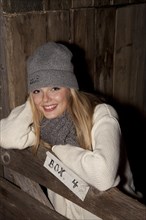 Smiling young woman wearing a hat and scarf standing at the gate of a horse's stall