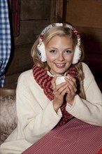Smiling young woman wearing ear warmers and a scarf sitting at the table of a hiking hut and drinking a hot drink