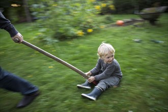 A little boy sitting on a shovel is dragged by an adult across the garden