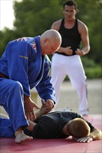 A martial arts instructor is demonstrating a holding grip with a student on an exercise mat in a park
