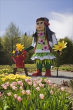 A tall native girl like doll stands next to a patch of pink and yellow tulips in the Greeting garden at the "Route des Gerbes d'Angelica" garden at springtime