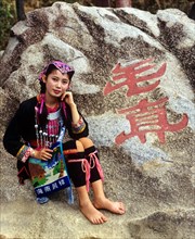 Young Miao woman wearing a traditional ethnic costume in front of rocks with a Chinese inscription