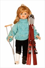 Doll with ski clothing and skis with her foot in plaster and holding a crutch following a ski accident