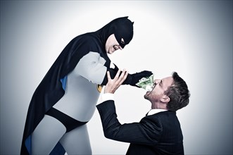 Chubby superhero wearing a Batman costume stuffing money into the mouth of a man wearing a suit