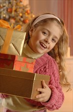 Smiling girl holding several gifts in front of a Christmas tree