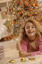 Smiling girl lying on the ground with a golden star and gifts in front of a Christmas tree