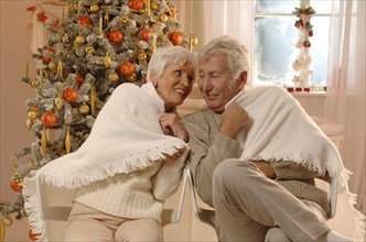 Mature couple in love wrapped in a blanked sitting in front of a Christmas tree