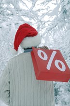 Man wearing a Santa hat in a snow-covered forest holding a shopping bag with a percent sign