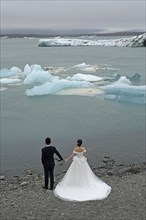 Bride in a classic white wedding dress and groom in a dark suit looking out over floating blocks of ice and the glacier on the banks of the Jokulsarlon lagoon
